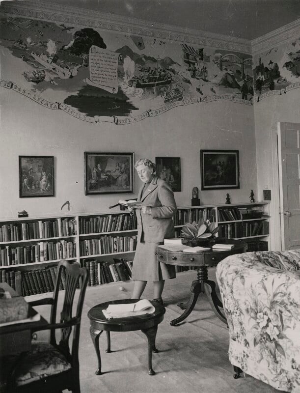 black and white photograph of Agatha standing in the library at Greenway beneath the mural painted by Lee Marshall.
