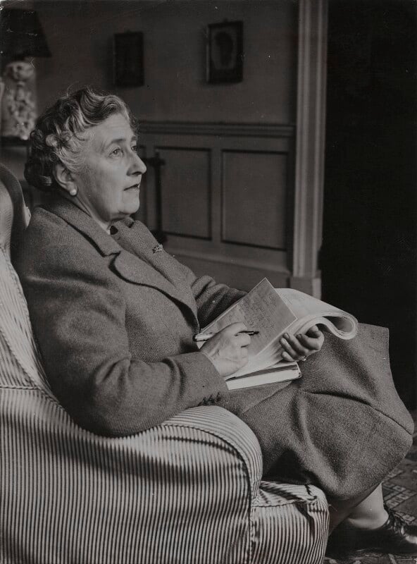 black and white photograph of Agatha sitting in a chair at Greenway with a notebook and pen in her lap.